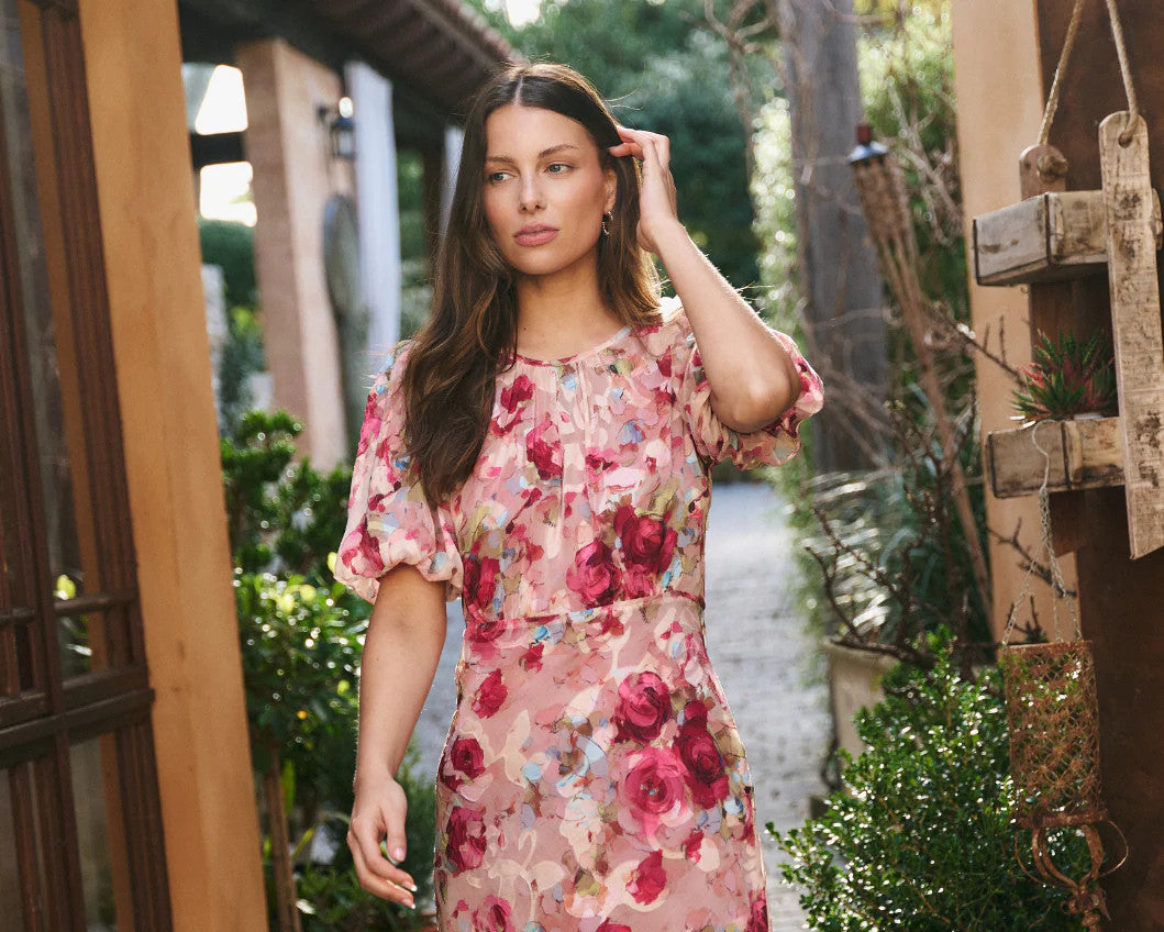 Woman in a floral dress walking on a stone path with plants and wooden structures around.